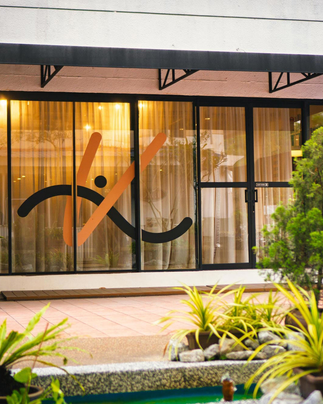 Building facade with a logo on the glass door, surrounded by plants.